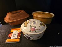 Photo showing Romertopf clay baker, Royal Worcester casserole with lid decorated with fruit and vegetable designs, and large mustard yellow oval casserole with lid along with Romertopf cooking booklet.