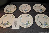 Six 6.5-inch plates in white with blue flowers and green leaves, gold rim, arranged on black surface