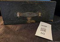 Closed view of the black carrying case with leather handle and metal clasp, showing wear and tear.