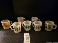 Photo showing all seven vintage milk glass mugs with various graphic patterns on a dark background.