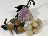 Photo showing all three fruit clusters together on a white background: purple amethyst grapes, white polished stone grapes, and brown carved stone or resin fruits with metal leaves and stems.