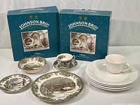 Two boxed 5-piece place settings of Johnson Bros Friendly Village dinnerware with one set unpacked showing plates, bowl, cup, and saucer; also four Pfaltzgraff plates and one coffee cup with saucer.