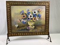 Front view of vintage fireplace screen showing floral print in ornate pressed tin frame with scrolled legs.