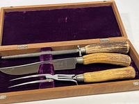 Top-down view of three-piece carving set inside wood box with purple velvet lining showing knife, fork, and sharpening steel with wood handles.