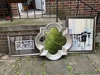 Two silver framed art pieces and one silver decorative mirror displayed outside on pavement. One art piece is a black and white floral photo, another a color painting of people with umbrellas, and a quatrefoil shaped mirror in silver frame.