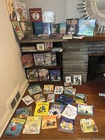 Wide shot showing many children's books arranged on shelves and floor, including board, hardcover, and paperback books. Titles such as 'Curious George,' 'Olivia,' and dinosaur books visible.