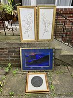 Full view of all four framed art pieces displayed outdoors on steps and a ledge. Two botanical line drawing pieces in gold frames are positioned at the top, a large blue abstract framed piece below, and a small square framed bronze medallion relief at the bottom.