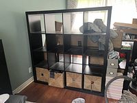 Front view of the black IKEA KALLAX shelf unit showing 16 square compartments with some woven baskets inside (not included). Room background visible.
