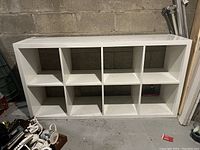 Full view of IKEA KALLAX white shelving unit with 8 compartments, placed against a basement wall. Visible faint scratches on top surface. Good used condition.