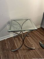 Full view of the square accent table with a metal base and clear glass top on wood floor next to wall.
