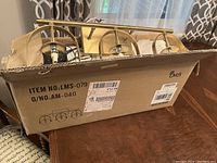 Gold three-light bathroom vanity light fixture partially inside original LMS branded box with shipping labels.