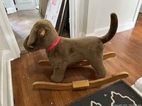 Side and partial rear view of the plush rocking dog showing brown fabric dog with raised tail, red collar, and wooden rockers on hardwood flooring.