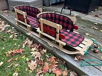 Twin wooden sleigh on deck and adjacent grass with red and black buffalo plaid cushions and green pull rope