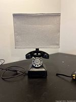 Front view of black telephone-shaped base table lamp with rotary dial detail and attached rectangular fabric shade on top.