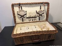 Open picnic basket showing intact wicker, interior fabric lining, leather straps, accompanying melamine dishware including plates, cups, and knives fixed in place