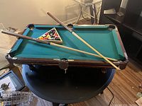 Full view of the miniature tabletop pool table set on a round table showing felt surface, balls in triangle rack, and two cues across the table.