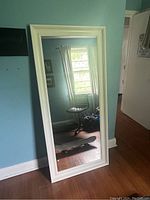 Full front view of the white framed full-length mirror leaning against blue wall, showing reflection of room and skateboard on floor.