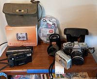 Photo showing the full lot including Canon digital camera with box, Canon AE-1 film camera with lens, Minolta film camera, Kodak Instamatic, Fujifilm cameras, extra lenses, and green carry case.