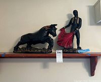 Photo showing the ceramic matador holding a red blanket and the bull sculpture side by side on a wooden shelf.