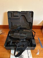 Hitachi camcorder inside open black hard plastic carrying case with strap and various cables including power adapter visible. Instruction manual partially visible beneath the case.