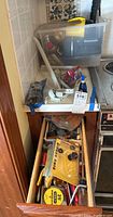 Photo of kitchen counter with plastic bin on it filled with various small tools, hardware, and a yellow handled screwdriver alongside a white painted curtain bracket and red handled tools.