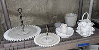 Overview of six assorted white milk glass items on a shelf against concrete wall: two tiered serving trays with metal handles featuring ruffled edges, a grape pattern covered dish, a hobnail bowl, a scalloped basket with handle, and a small round dish.
