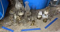 Overview of 12 vintage oil lamps with chimneys and bases arranged on floor near plastic bin.