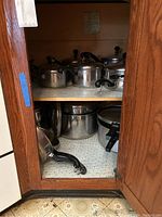 Cabinet shelf showing multiple stainless steel pots and pans of various sizes, all with black plastic handles.