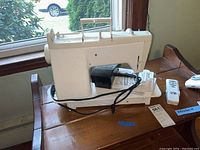 Side and top view of tailor sewing machine on wooden surface with power cord and user manual underneath.