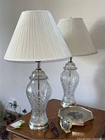Two glass and metal table lamps with pleated white lampshades, an octagonal wall clock and a small round alarm clock on wooden table.