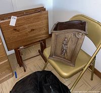 Photo showing three medium-toned wooden TV trays with foldable stands, a beige folding chair, and the decorative waste basket arranged together.