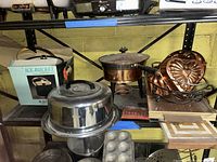 Photo of vintage cake storage items on basement metal shelf, showing stainless steel cake dome, fondue pot, copper heart-shaped cake mold on stand, and boxed black octagonal ice bucket.