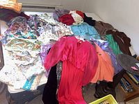 Lot of assorted vintage clothing spread out including a prominent bright pink dress with sheer sleeves and other shirts, sweaters, and jackets in various colors and styles.