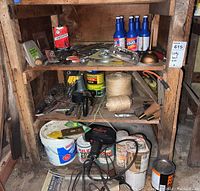 Wooden shelf with various vintage hand tools, clamps, cans of automotive maintenance products, paint, coiled string, and electric drill