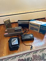 Photo showing all 5 items included in the lot laid out on a wooden floor: Panasonic RF-559 radio with box, GE digital clock radio cassette player, RCA Victoria radio, Mondale AM radio, and Sony Walkman with carrying pouch.