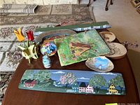 Overview of various decorative wooden and painted items on a table including painted panels, carved tulips, shelf, plate, and other art pieces.