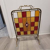 Front view of brass frame stained glass fireplace screen showing multicolored panels and decorative handle, note missing and broken glass pieces.