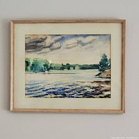 Framed watercolour painting depicting a body of water with trees and cloudy sky in soft muted tones, mounted on wall.