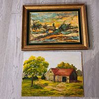 Both oil paintings placed on floor side by side. Top painting is abstract landscape with sunset framed in gold frame, bottom painting shows barn and tree on canvas unframed.