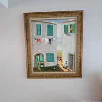 Frontal view of framed painting depicting Mediterranean village facade with green shutters, laundry line and figures
