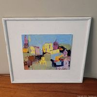 Framed painting showing the colorful cityscape with its white wooden frame against a light background.