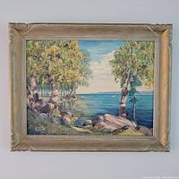 Framed oil on board landscape painting showing trees, lake, cloudy sky, rocks, and grass.