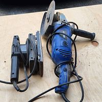 Photo showing blue circular saw and black belt sander lying on wooden surface from top angle.