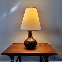 Vintage ceramic table lamp with dark brown glossy base and cream tapered shade on wooden table, illuminated to show light effect