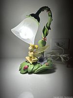 Full view of the lamp lit, showing Tinkerbell sitting on the lily pad base with flowers and a ladybug, and the frosted bellflower glass shade illuminated.