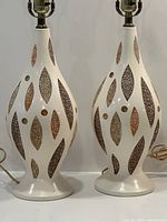 Front view of both lamps showing off-white ceramic bases with elongated teardrop silhouette and brown/tan textured inserts in leaf-shaped reliefs.