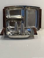 Two stainless steel serving trays with teak handles; one tray has three compartments, the other has one. A pair of conical stainless steel salt and pepper shakers with patterned bands are placed in the smaller tray.