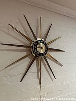 Full view of the Ingraham Mid-Century Modern starburst wall clock hanging on a white brick wall, showing the radial wooden and metal spokes and central clock face.