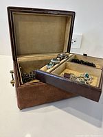 Open brown suede jewelry box showing interior with assortment of costume jewelry inside including bracelets and rings.