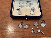 Overview of all cufflinks in the set placed on a wooden surface showing the collection with the velvet box containing the largest pairs behind.